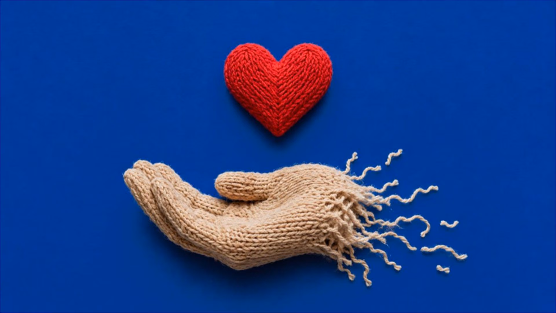 A knitted red heart resting above a knitted hand, unravelling at the wrist