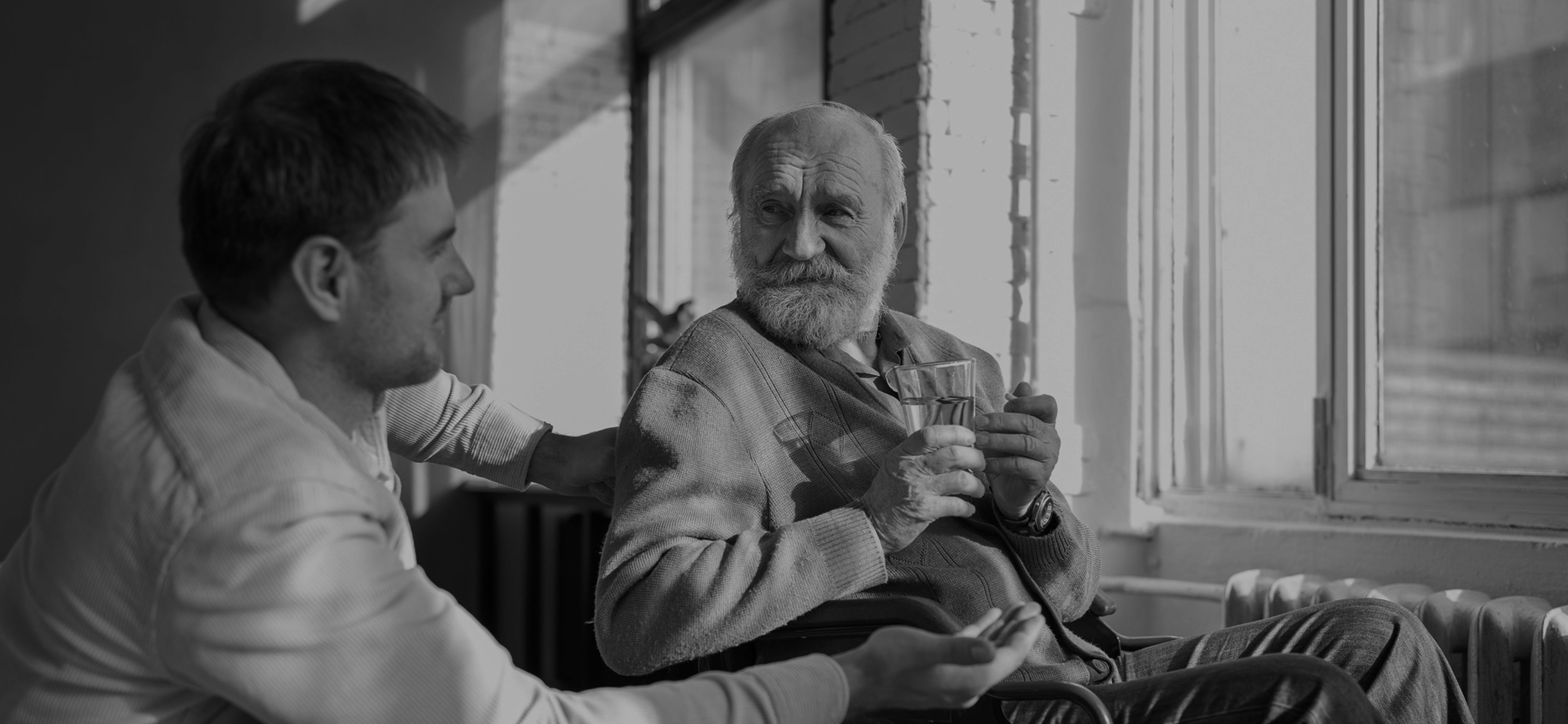 A young man crouching beside an older man in a wheelchair, who holds a glass of water by a sunlit window.