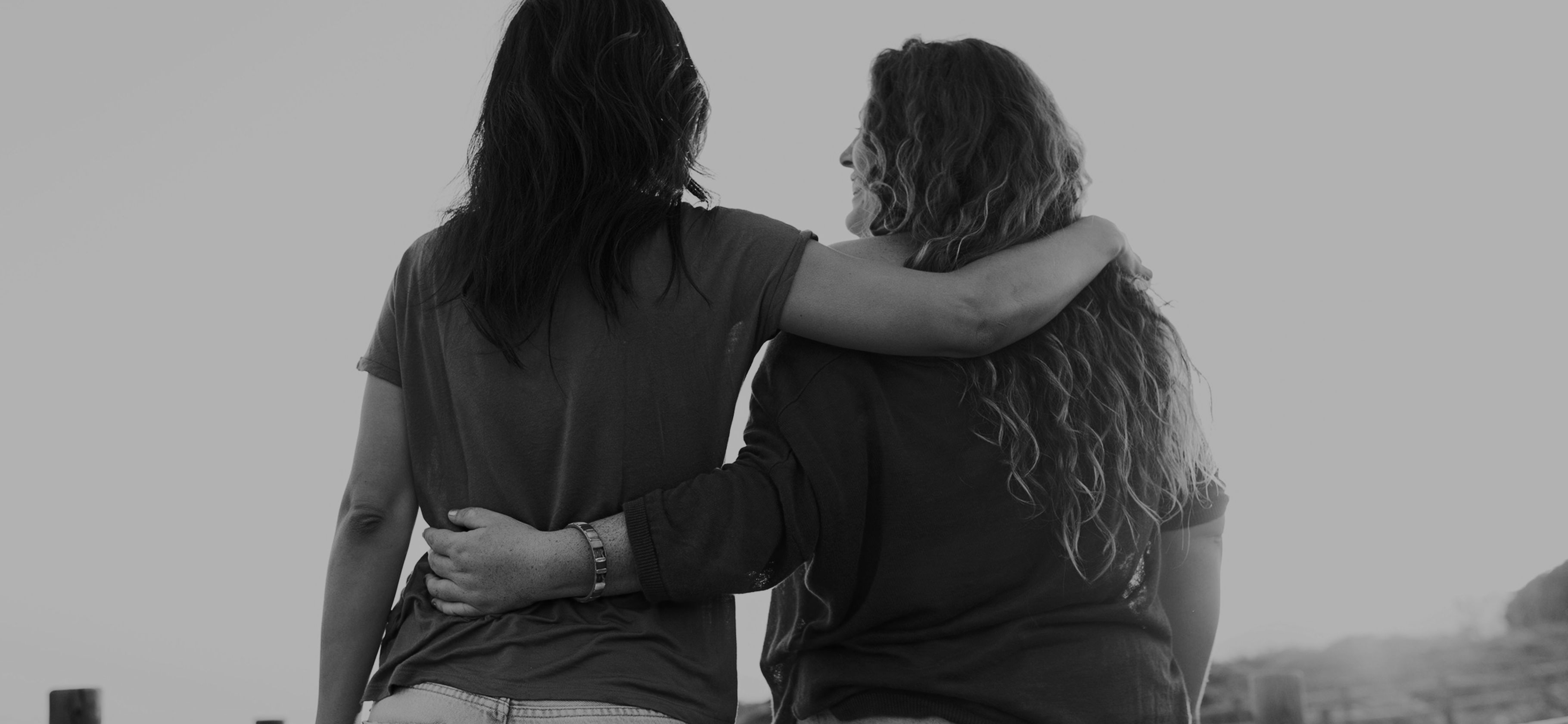 Two women walking away along a boardwalk at sunset, arms around each other.